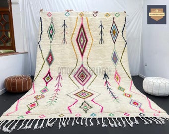 May include: A white Moroccan rug with a geometric pattern in shades of pink, green, blue, and yellow. The rug has a fringe border and is made of wool.