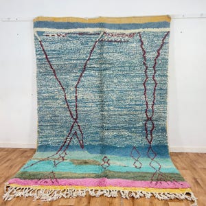 May include: A large, rectangular rug with a textured, woven design. The rug features a gradient of colors, including blue, turquoise, pink, and yellow. Dark red geometric lines and shapes are woven into the blue field. The rug has a fringe along the bottom edge.