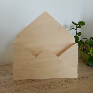 Könnte beinhalten: Ein hölzerner Wandorganizer in Form eines Umschlags mit einer hellen Holzoptik. Der Organizer hat zwei messingfarbene Haken am oberen Klappen.