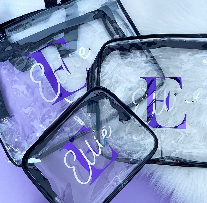 May include: Three clear plastic zippered bags with black trim. The bags are personalized with the name "Ellie" in a white cursive font and a purple letter "E".