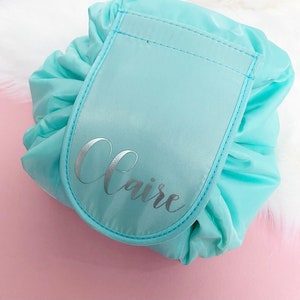 May include: A light blue drawstring makeup bag with a large pocket on the front. The pocket is also light blue and has the name "Claire" written in silver lettering.