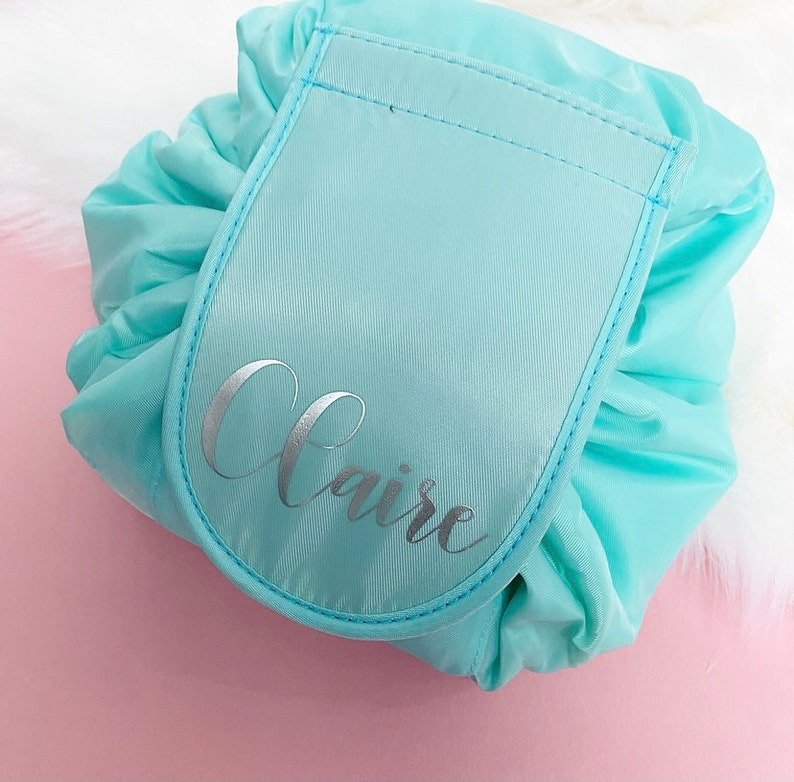 May include: A light blue drawstring makeup bag with a large pocket on the front. The pocket is also light blue and has the name "Claire" written in silver lettering.