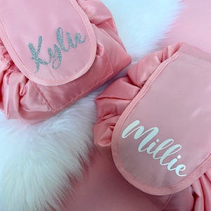 May include: Two pink drawstring makeup bags with personalised names. The bag on the left says "Kylie" in silver glitter lettering. The bag on the right says "Millie" in white lettering.