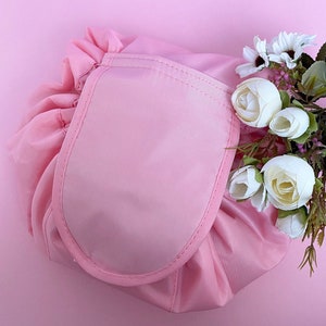 May include: A pink makeup bag with a large, round, pink pocket on the front. The bag is made of a soft, silky material and has a drawstring closure. The bag is surrounded by white flowers.