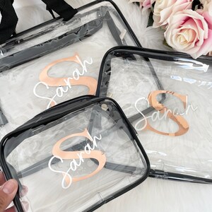 May include: Three clear plastic zippered travel bags with black trim. Each bag has a gold letter 'S' and the name 'Sarah' written in white on the front.