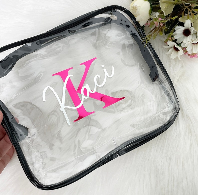 May include: Clear plastic zippered pouch with a black zipper and trim. The pouch has a pink letter "K" and the name "Kaci" written in white cursive.