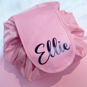 May include: A pink drawstring makeup bag with a large flap that has the name "Ellie" written in black cursive lettering.