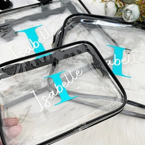 May include: Three clear plastic zippered bags with black trim. The bags are personalized with the name "Isabelle" and the letter "I" in turquoise.