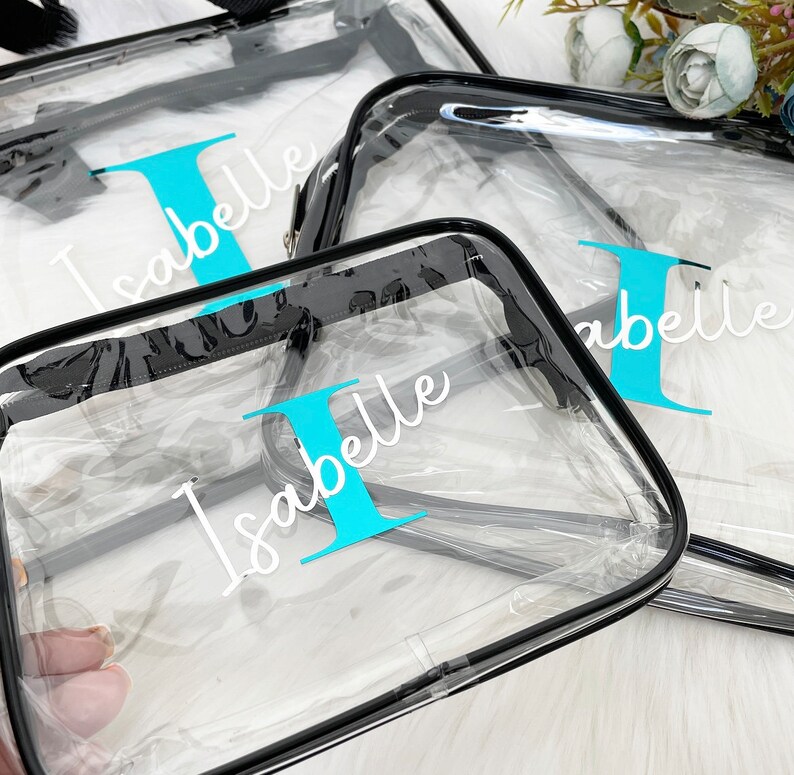 May include: Three clear plastic zippered bags with black trim. The bags are personalized with the name "Isabelle" and the letter "I" in turquoise.