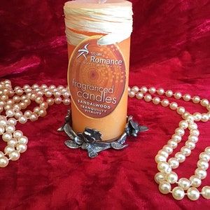 May include: A round orange candle with a label that reads "Mount Romance Uniquely Australian Fragranced Candles Sandalwood Tranquility Vitality" is sitting on a silver metal candle holder with a floral design. The candle is surrounded by a string of white pearls on a red velvet surface.