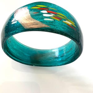 May include: Teal-colored glass bangle bracelet with a decorative design. The bracelet features a gold-colored accent and colorful streaks of red, yellow, and white. The bangle is a solid, rounded shape.
