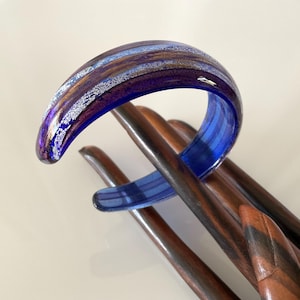 May include: A striking glass bangle in blue, purple, and silver tones is displayed on dark brown wooden rods. The bracelet has a layered colour design and a glossy finish, creating a contrast.