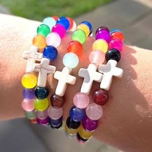 May include: A collection of colorful beaded bracelets. Each bracelet features a mix of vibrant, round beads in various colors, including pink, blue, green, and yellow. White cross-shaped charms are interspersed throughout the design, creating a religious theme.