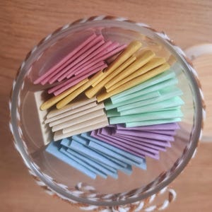 Op de afbeelding: Een heldere glazen pot met een rand van gevlochten touw, gevuld met stapels gekleurde papieren briefjes in roze, geel, groen, crème, paars en blauw. De briefjes zijn in nette rijen gerangschikt.