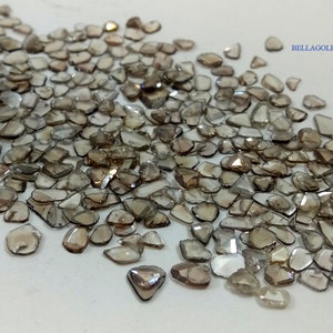 May include: A large collection of rough, unpolished, brown diamonds. The diamonds are of various shapes and sizes, and they are scattered on a white surface.  BELLAGOLDGEMS is printed in the upper right corner of the image.