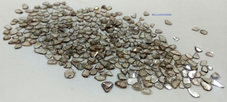 May include: A large collection of rough, unpolished, brown diamonds. The diamonds are of various shapes and sizes, and they are scattered on a white surface.  BELLAGOLDGEMS is printed in the upper right corner of the image.