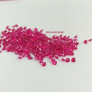 Puede incluir: Un montón grande de piedras preciosas de rubí rosa brillante sueltas. Las piedras preciosas son redondas y facetadas. Las piedras preciosas están dispersas sobre una superficie blanca. BELLAGOLDGEMS está escrito en texto rojo en la imagen.