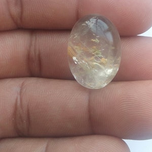 以下が含まれることがあります： 金色と緑色のインクルージョンが入った透明な楕円形の宝石。宝石は手で持たれています。