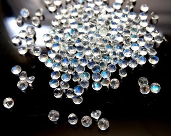 May include: A pile of iridescent white moonstone cabochons, each with a unique blue sheen. The stones are round and smooth, and they are scattered on a black surface.
