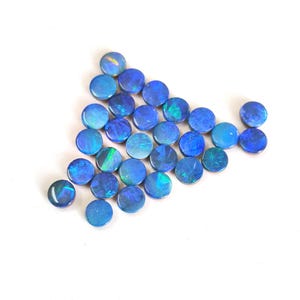 May include: A collection of round, flat-backed cabochons featuring a deep blue base colour with iridescent flashes of green and light blue light. These small, circular gemstones are arranged in a loose triangular formation on a plain surface.