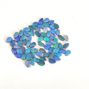 May include: A collection of numerous small, teardrop-shaped loose opals. Each gemstone features a deep blue base colour with iridescent flashes of bright green and lighter blue light reflecting from within the polished, smooth surfaces.