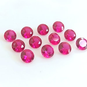 May include: A group of twelve round, faceted, ruby red gemstones. The gemstones are scattered on a white surface.