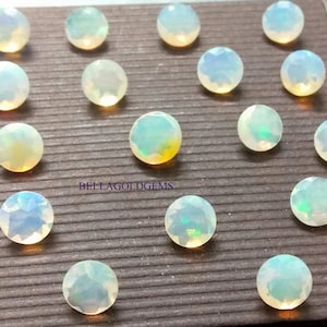 Può includere: Una collezione di 16 pietre preziose rotonde di opale bianco con un leggero bagliore iridescente. Le pietre preziose sono disposte in file su una superficie marrone testurizzata. Il testo "BELLAGOLDGEMS" è visibile al centro dell'immagine.