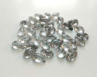 May include: A collection of pear-shaped, clear quartz gemstones with black inclusions. The stones are arranged in a loose pile on a white surface.