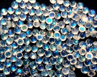 May include: A collection of round, iridescent moonstone cabochons in various shades of blue and white. The stones are arranged in a loose pile on a black background.