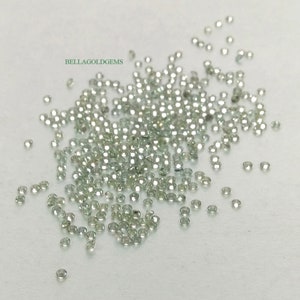 May include: A loose pile of small, round, clear green gemstones. The gemstones are scattered on a white surface. The text "BELLAGOLDGEMS" is visible in the upper left corner of the image.