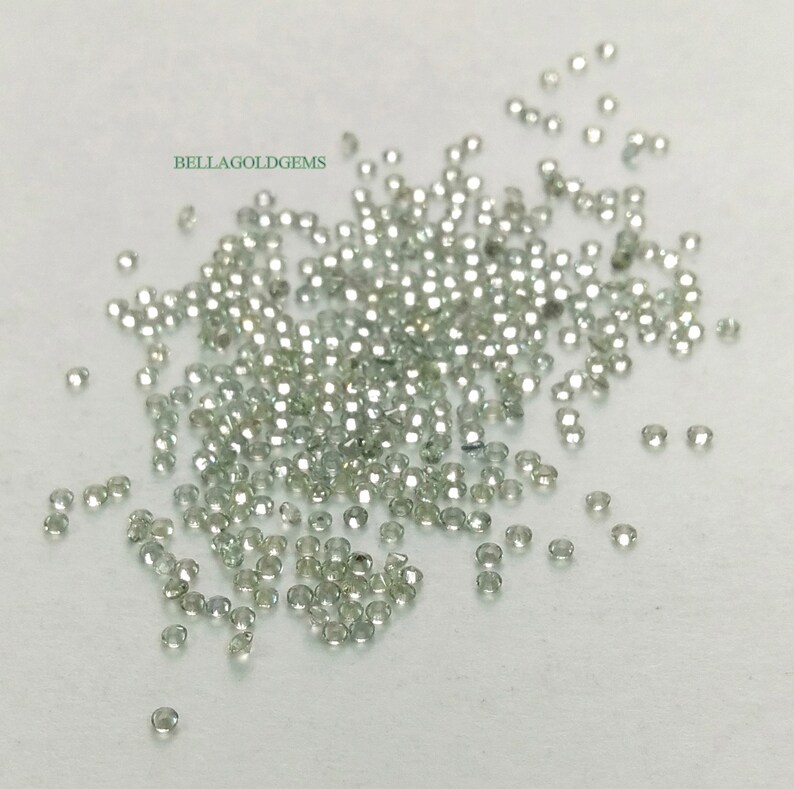 May include: A loose pile of small, round, clear green gemstones. The gemstones are scattered on a white surface. The text "BELLAGOLDGEMS" is visible in the upper left corner of the image.