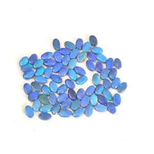 May include: A collection of numerous small, oval-shaped loose gemstones. Each stone features a deep blue base colour with iridescent flashes of bright cyan and green light reflecting across the polished, smooth surfaces of the stones.