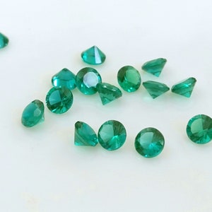 May include: A collection of round, faceted, green gemstones. The gemstones are scattered on a white surface.