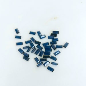 Op de afbeelding: Een verzameling donkerblauwe rechthoekige edelstenen, waarschijnlijk saffieren, verspreid over een wit oppervlak.