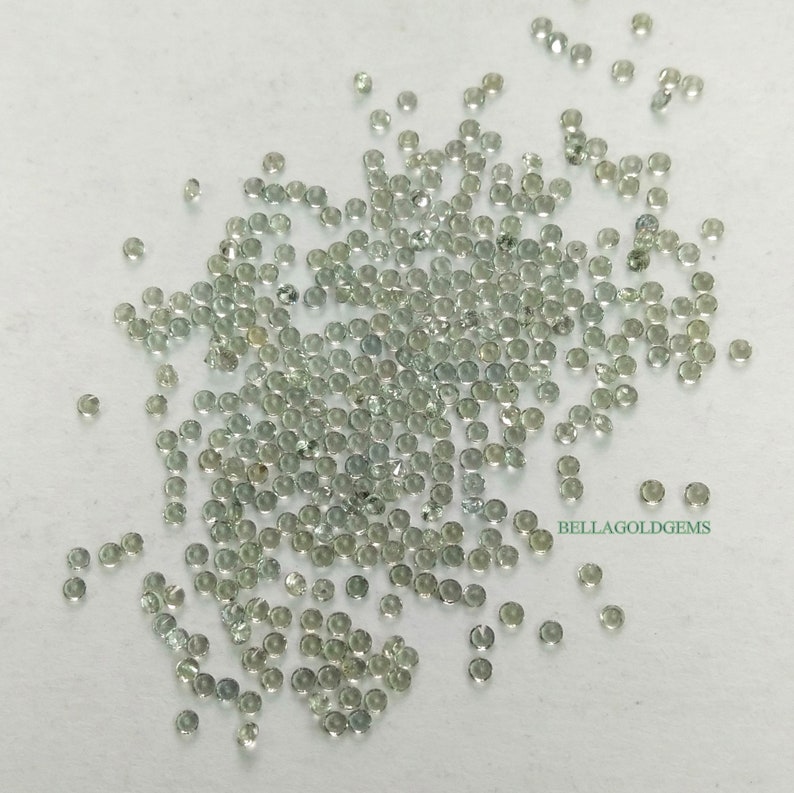 May include: A large quantity of small, round, light green gemstones. The gemstones are scattered on a white surface.  BELLAGOLDGEMS is printed in the lower right corner of the image.