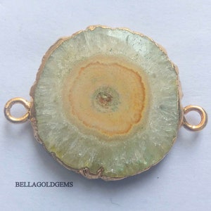 May include: A round, light green and brown agate slice with gold-toned metal accents. The agate has a circular pattern of light brown and white. The slice is attached to two gold-toned metal loops.