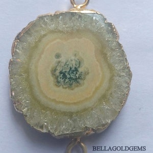 May include: A round, light green and white agate slice with a gold-toned metal connector on each side. The agate has a natural, crystal-like texture.  BELLAGOLDGEMS is printed on the bottom right corner of the image.