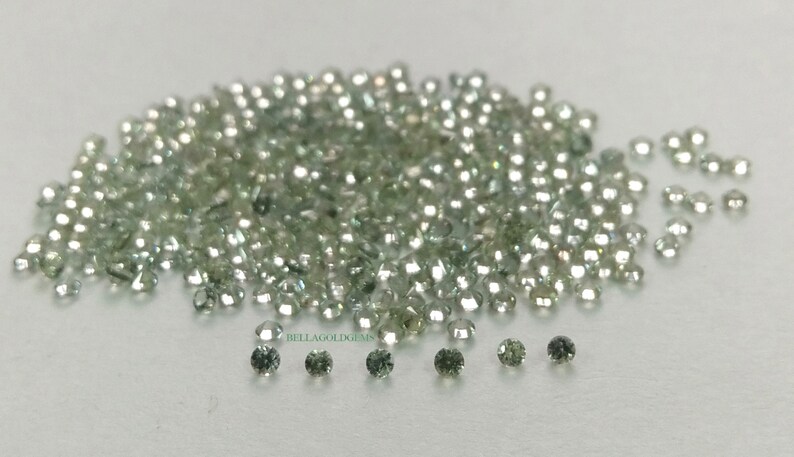 May include: A large pile of small, round, green and clear gemstones. The gemstones are scattered on a white surface.