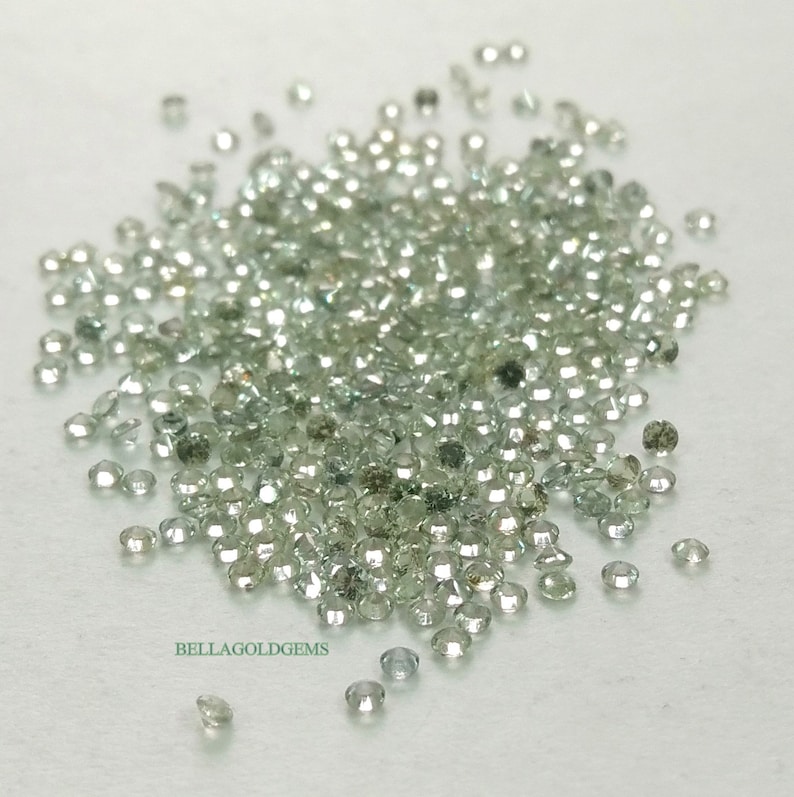 May include: A pile of small, round, light green gemstones. The gemstones are scattered on a white surface. The gemstones are all about the same size and shape.