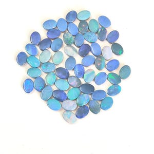 May include: A collection of numerous oval-shaped blue opals arranged in a cluster. Each gemstone displays a smooth, polished surface with varying shades of blue, ranging from light sky blue to deep navy, featuring iridescent flashes of green and white.