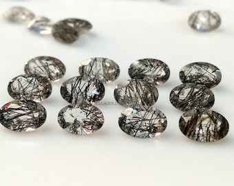 May include: A collection of oval-shaped, clear quartz gemstones with black inclusions. The stones are arranged in a row on a white surface.