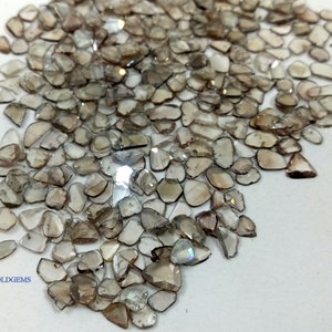 May include: A large pile of small, irregular, light brown, faceted gemstones. The gemstones are scattered on a white surface. The text "BELLAGOLDGEMS" is visible in the lower left corner of the image.