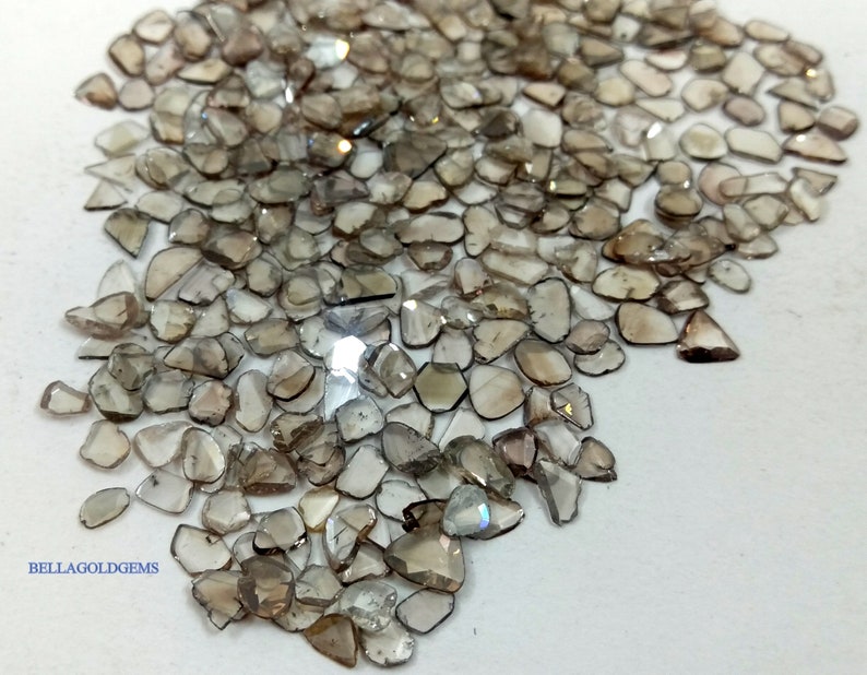 May include: A large pile of small, irregular, light brown, faceted gemstones. The gemstones are scattered on a white surface. The text "BELLAGOLDGEMS" is visible in the lower left corner of the image.