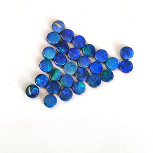 May include: A collection of small, circular, polished blue opals arranged in a triangular formation. Each gemstone displays a deep blue base colour with iridescent flashes of green and light blue light reflecting across the smooth, rounded surfaces.