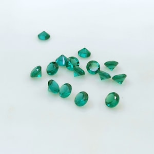 May include: A collection of small, faceted emerald-green gemstones scattered on a white surface. The gemstones vary in shape, with some appearing round and others more pointed. The light reflects off the facets, creating a sparkling effect. These could be used for jewelry making or other crafts.