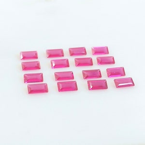Puede incluir: Una colección de 15 piedras preciosas rectangulares de color rosa, probablemente rubíes, dispuestas en un patrón escalonado sobre una superficie blanca.