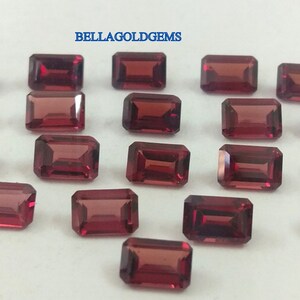 May include: Twenty-two loose, faceted, rectangular, red gemstones. The gemstones are arranged in a heart shape on a white surface. The text "BELLAGOLDGEMS" is printed in blue at the top of the image.