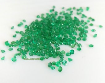 May include: A large pile of round, green emerald gemstones. The gemstones are scattered on a white surface.