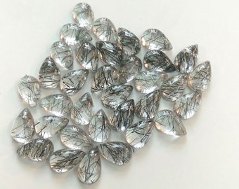 May include: A collection of clear, teardrop-shaped gemstones with black needle-like inclusions. The gemstones are scattered on a white surface, showcasing their transparency and unique internal patterns. These stones are likely used for jewelry making.