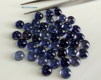 May include: A collection of round, polished, dark blue gemstones. The gems are scattered across a white surface, with a pair of tweezers in the background. The gemstones have a smooth, reflective surface.
